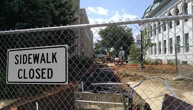 A sign on a fence reads, 'Sidewalk Closed,' with construction in the background